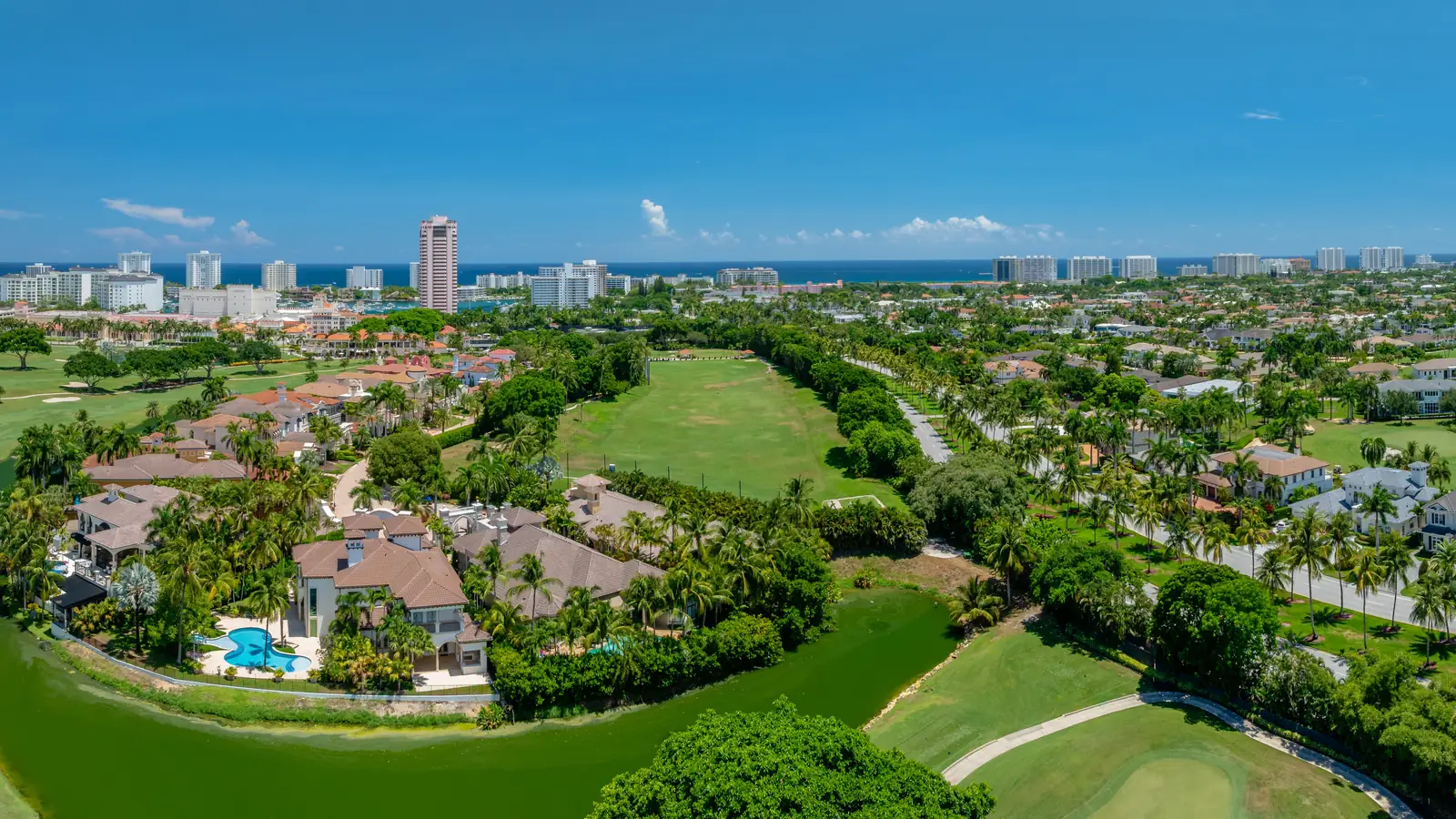 Aerial view of a South Florida golf community: barrel-tiled houses with swimming pools set among palms along a fairway, a palm-lined boulevard running through the middle distance, and a skyline of white coastal towers with the Atlantic Ocean behind.