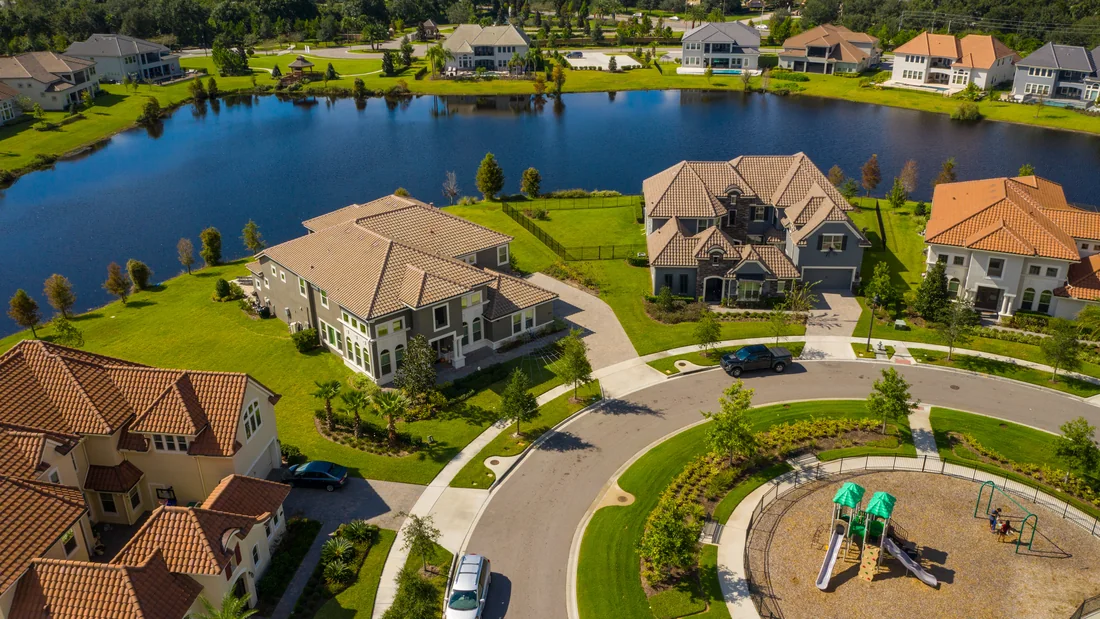 Aerial view of a suburban Florida lakefront neighbourhood: two-storey barrel-tiled houses on landscaped lots around a cul-de-sac, a dark blue lake behind them, and a fenced children's playground with green shade canopies and swings.