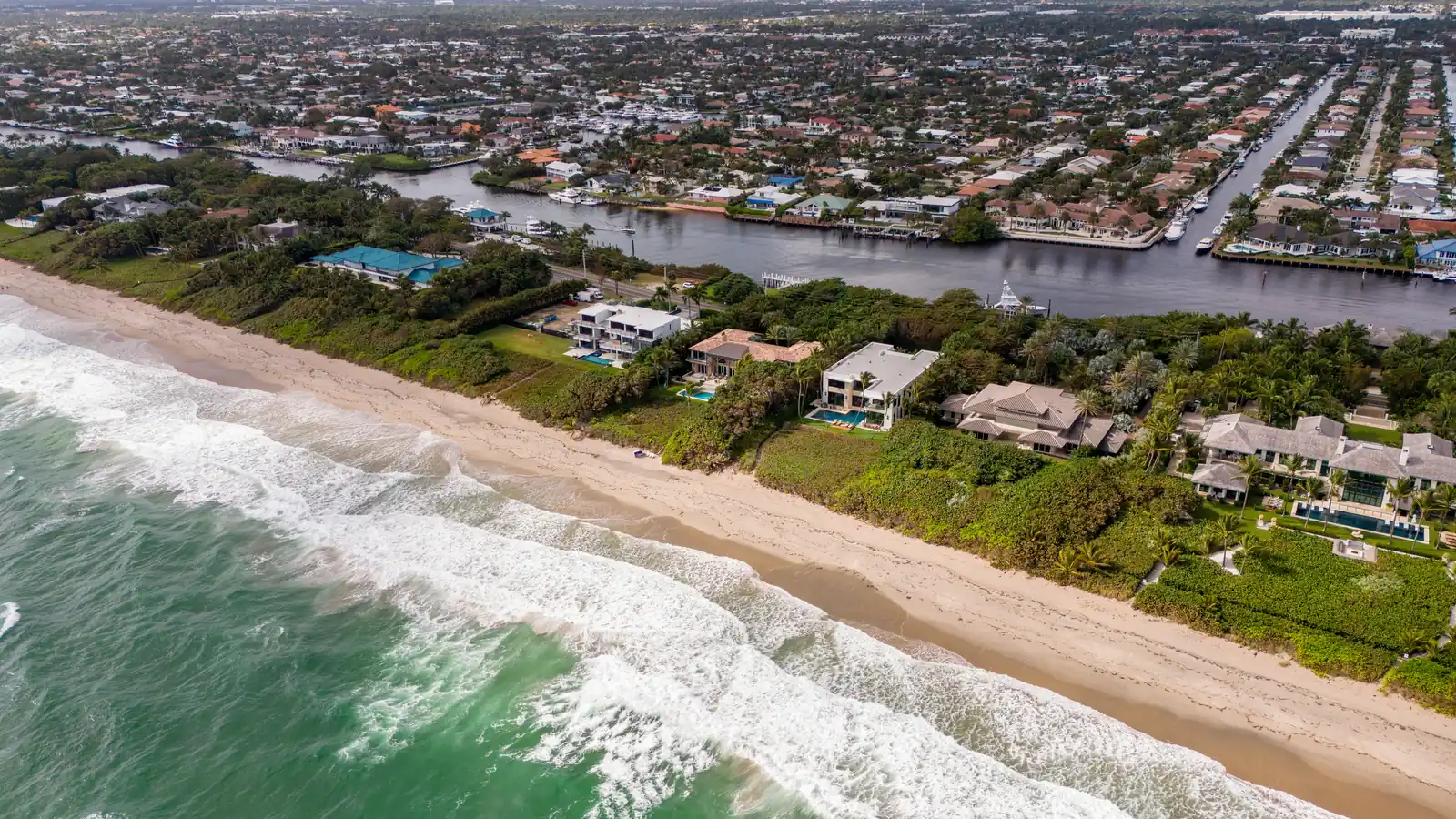 Aerial view of a South Florida coastline: lines of surf breaking onto a wide sand beach, oceanfront houses with swimming pools behind the dune line, and beyond them the Intracoastal Waterway with canal streets, private docks and moored boats.