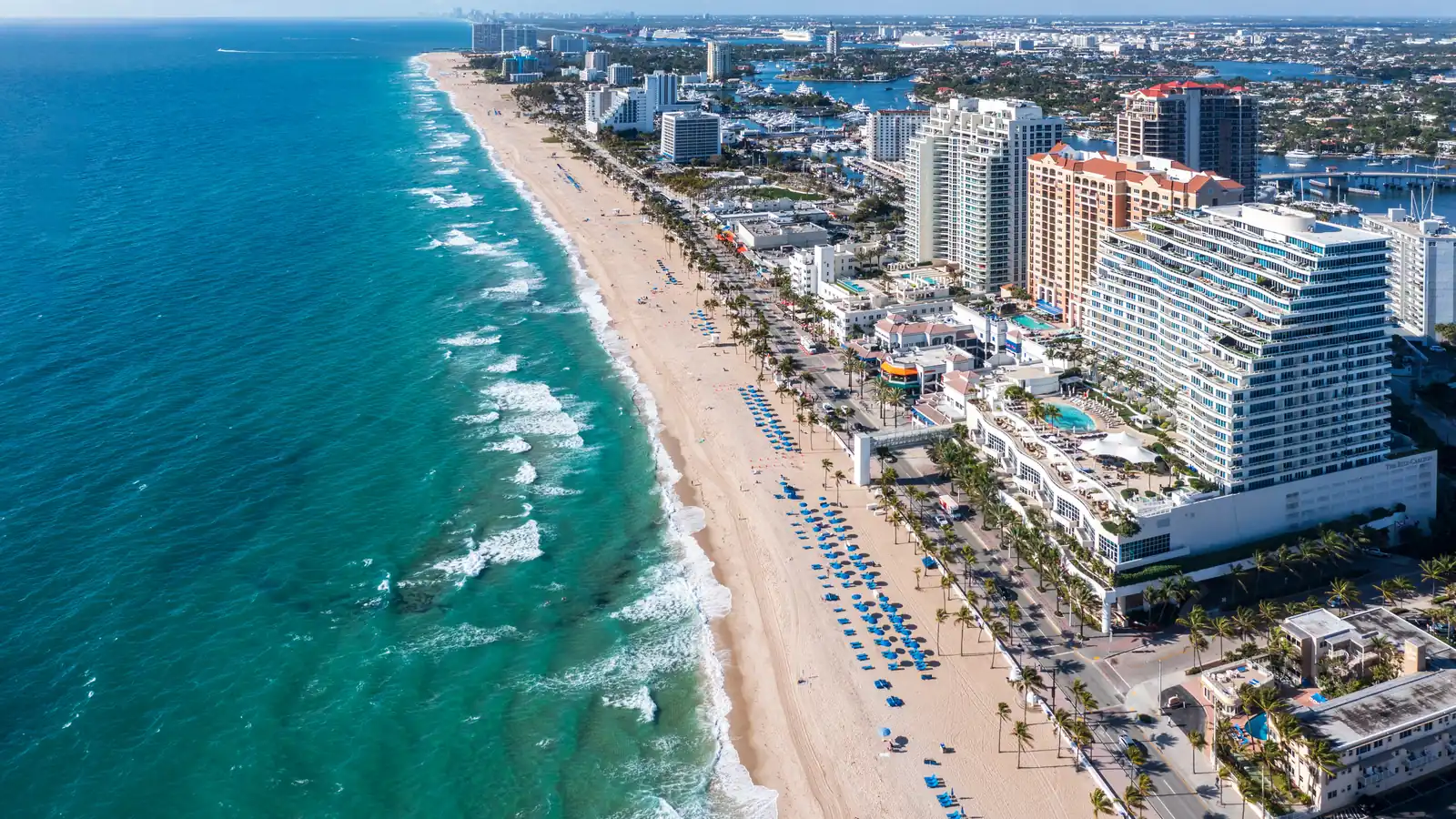 Aerial view along Fort Lauderdale Beach: a long straight wall of beachfront high-rise hotels and condos above wide sand, blue umbrellas in rows, surf along the shore, and the city stretching inland behind the towers.