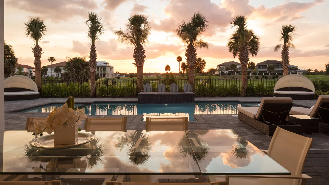 View at sunset from a covered lanai across a screened swimming pool: a glass outdoor dining table with orchids, wicker loungers and two round shaded daybeds, and beyond a low black fence a golf fairway lined with sabal palms and houses under a pink and orange sky.
