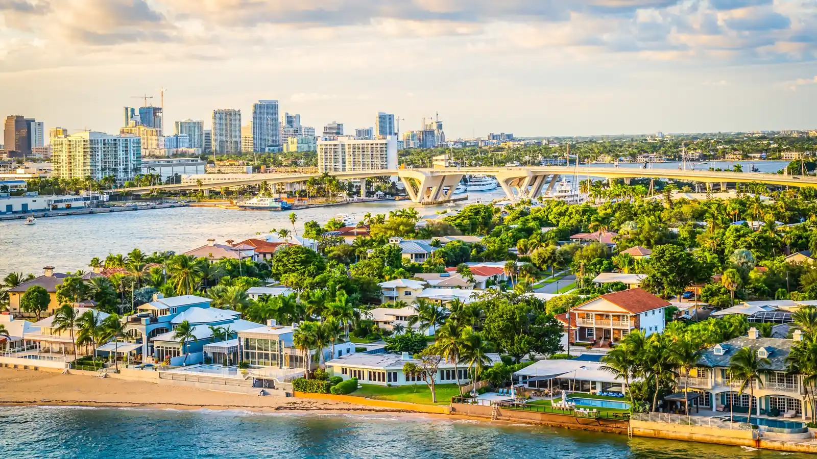 Aerial view at golden hour over Biscayne Bay in Miami: waterfront homes with pools along a residential island, a causeway bridge crossing the bay, and the downtown skyline rising on the horizon under warm evening light.