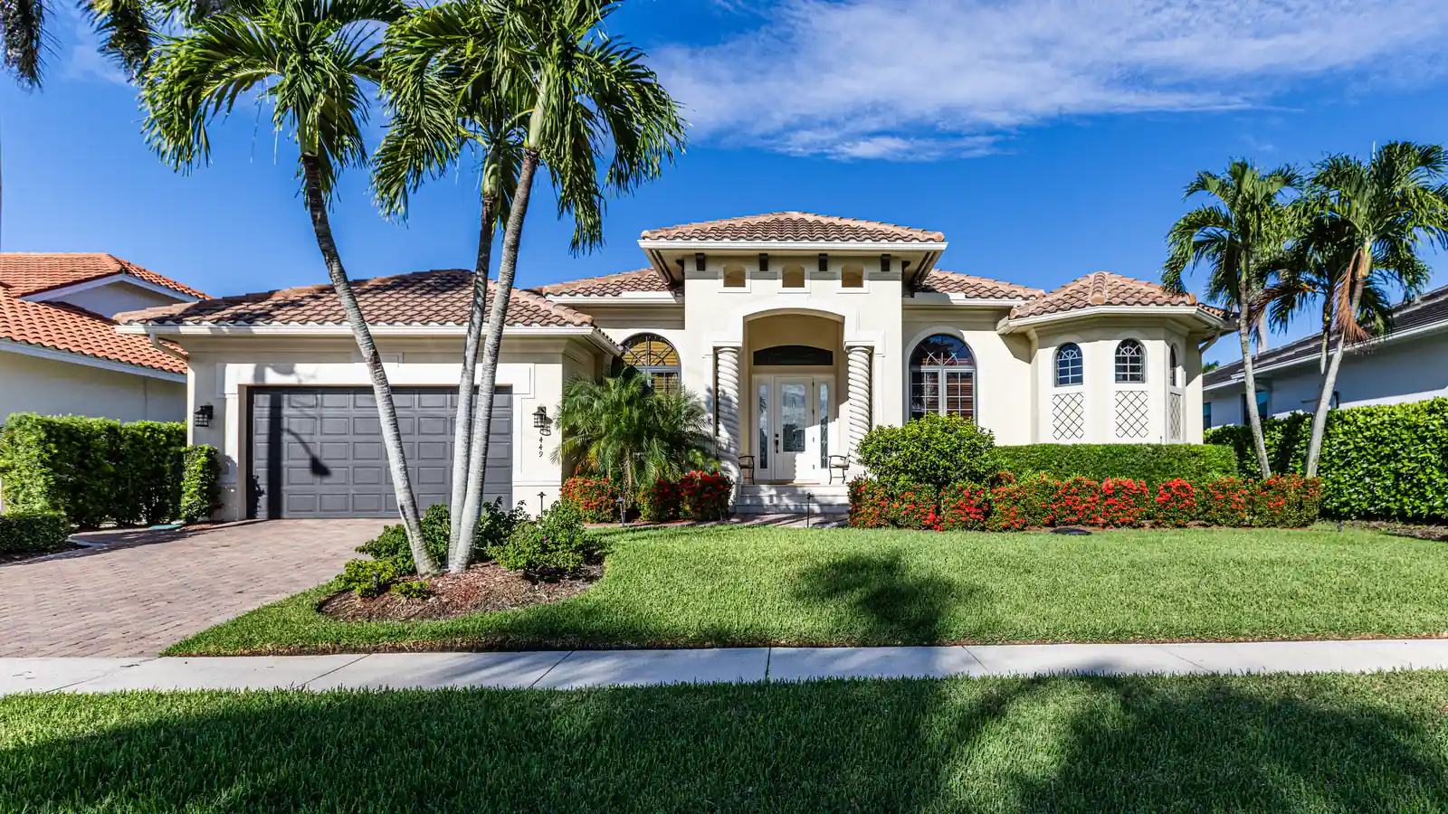 A single-storey cream Florida Mediterranean house with a barrel-tile roof, an arched columned entry and an arched bay window, royal palms on the lawn, a red flowering hedge along the foundation and a brick-paver driveway.