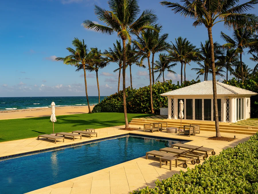 A beachfront lawn and long rectangular swimming pool with teak loungers and folded parasols, a white pool pavilion with a shingled hip roof and lattice panels, tall coconut palms and a sea-grape hedge, with sand and the Atlantic Ocean beyond.