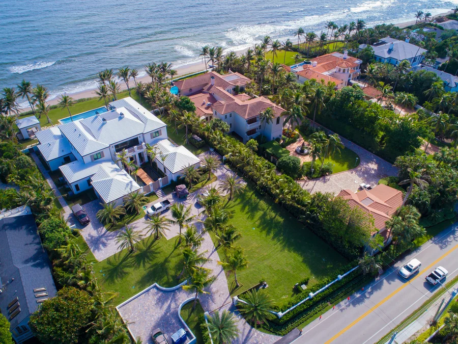 Aerial view of two large oceanfront properties side by side: one white with a standing-seam metal roof, one cream with a terracotta tile roof and a rounded tower, each with a swimming pool, paver motor court and clipped hedges, with coconut palms, a coastal road and the Atlantic surf beyond.