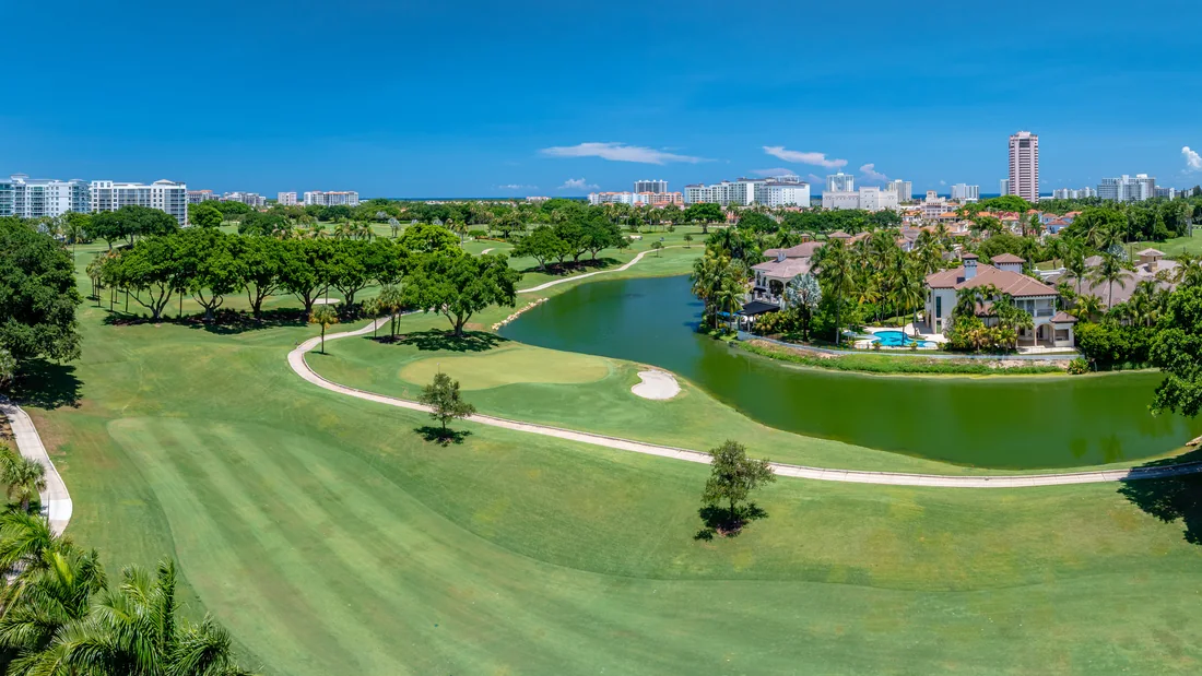 Aerial view over a South Florida golf community: a wide striped fairway and a putting green with a sand bunker in the foreground, a curving cart path, a lake, large tile-roofed homes with swimming pools along the far bank, and a line of coastal high-rise buildings on the horizon.
