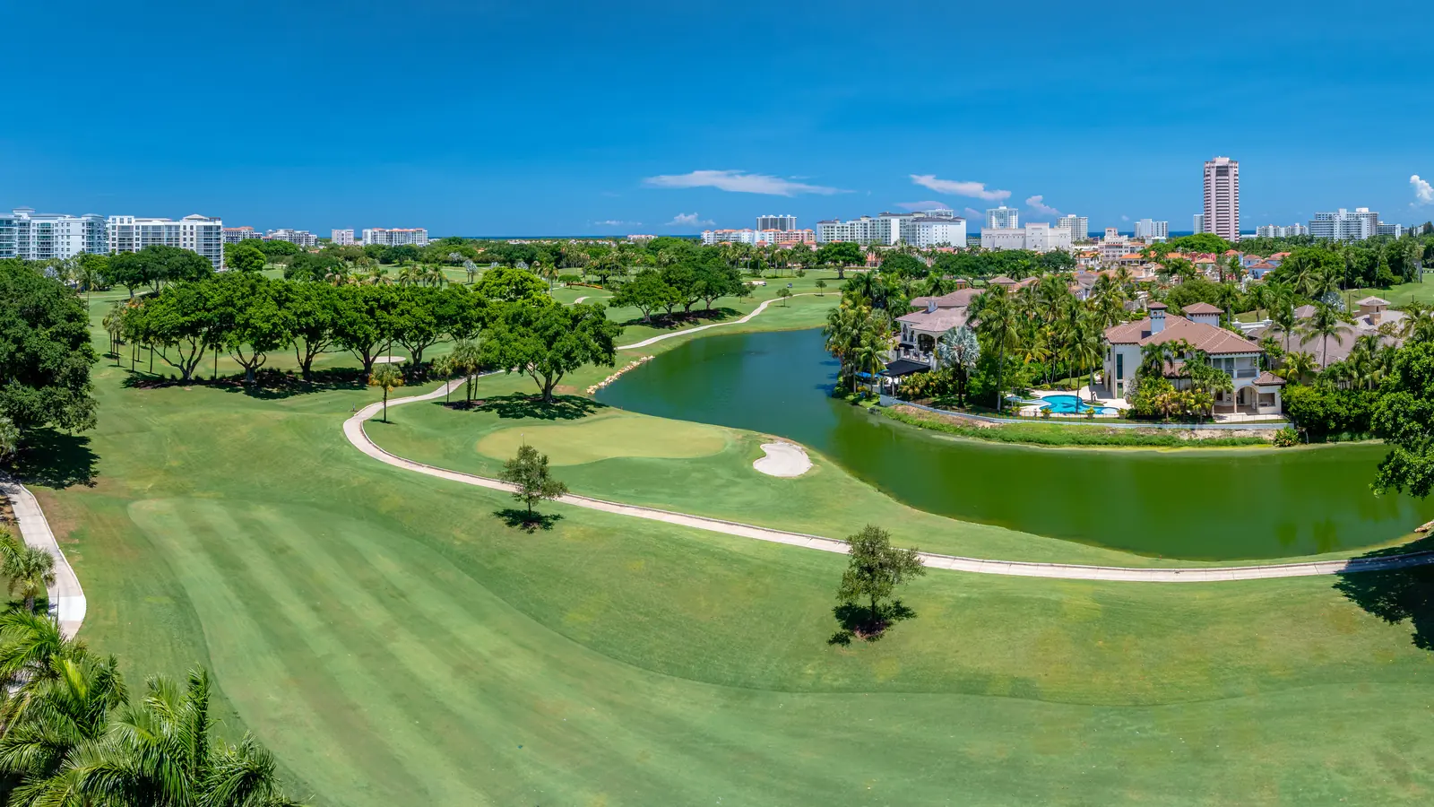 Aerial view over a South Florida golf community: a wide striped fairway and a putting green with a sand bunker in the foreground, a curving cart path, a lake, large tile-roofed homes with swimming pools along the far bank, and a line of coastal high-rise buildings on the horizon.