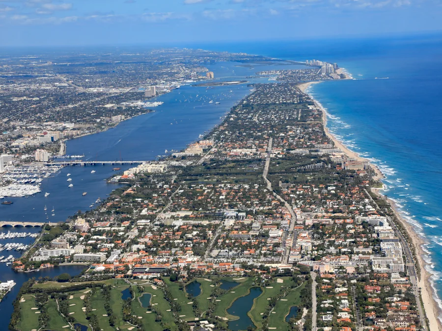 High-altitude aerial view of a South Florida barrier island: the Atlantic Ocean along one side, the Intracoastal Waterway along the other, and a narrow strip of green streets and rooftops running between them into the distance.