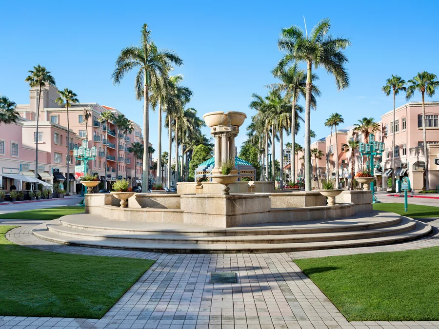 The stone fountain at the centre of Mizner Park in Boca Raton, framed by royal palms, pale pink Mediterranean-style buildings with shopfronts and balconies, and turquoise multi-globe lamp standards, under a clear blue sky.