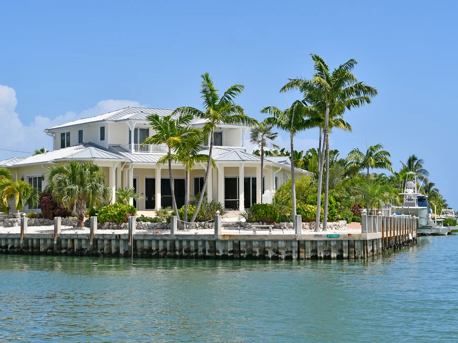A cream two-storey waterfront house with a metal roof and a deep covered porch, standing behind a concrete seawall on a turquoise canal, with coconut palms across the front lawn and a motor boat moored at the dock alongside.