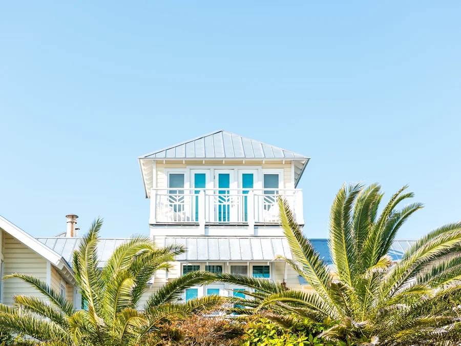 The upper storeys of a pale yellow coastal house with a standing-seam metal roof, a square rooftop lookout tower and a white balcony railing, seen over the fronds of date palms against a clear blue sky.