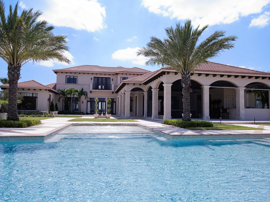 The rear elevation of a large Mediterranean-style house with a terracotta tile roof and an arched loggia on stone columns, seen across a long rectangular swimming pool, with date palms on the terrace under a blue sky with scattered cloud.