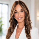 Kelly Scalone in a head-and-shoulders portrait: long layered brown hair with lighter highlights, a white blazer over a white top, small hoop earrings and a fine gold chain with a diamond cross pendant, photographed indoors beside a bright window.
