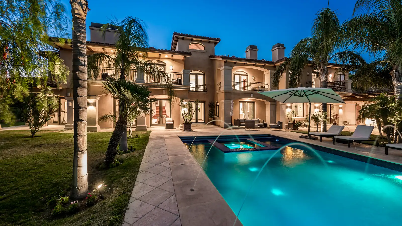 A two-storey Mediterranean-style house at dusk with warm interior lighting, arched loggias and wrought-iron balconies, seen across a lit swimming pool with arcing water jets, a spa, loungers and a parasol on the terrace.